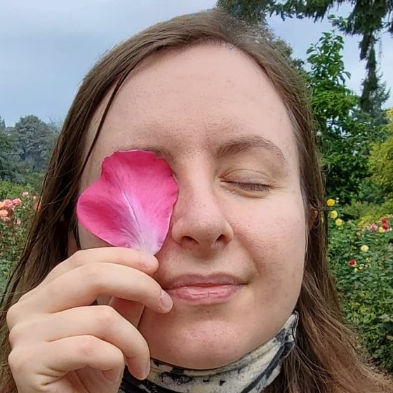 Maria Mishurenko holding a rose petal over one eye in a rose garden.